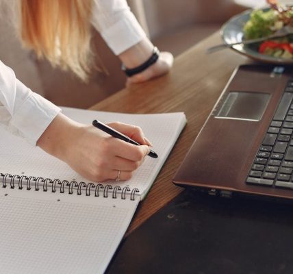 From above of crop anonymous female freelancer writing with pen in copybook while standing near table with laptop and plate of food for lunch in cafeteria