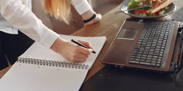 From above of crop anonymous female freelancer writing with pen in copybook while standing near table with laptop and plate of food for lunch in cafeteria