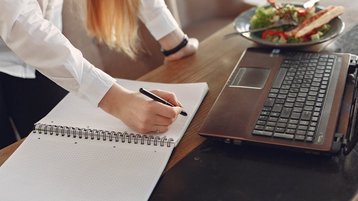 From above of crop anonymous female freelancer writing with pen in copybook while standing near table with laptop and plate of food for lunch in cafeteria